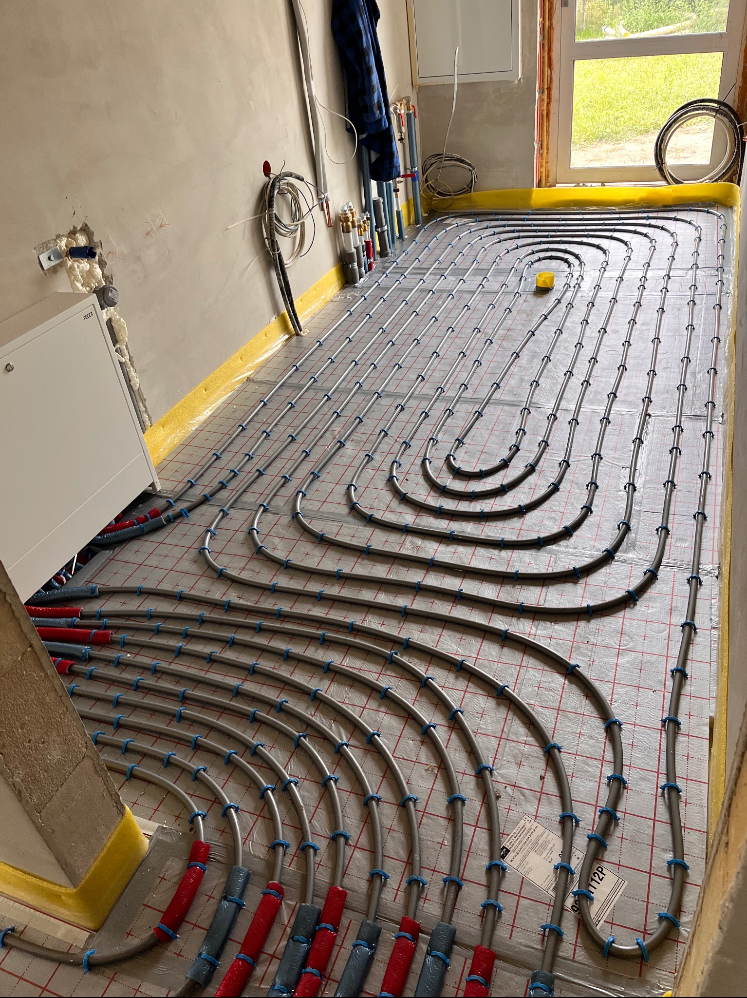 Szare rury ogrzewania podłogowego, przymocowane niebieskimi uchwytami do podłoża z siatką. Żółta taśma brzegowa wzdłuż ścian. Widok na okno i fragment instalacji.