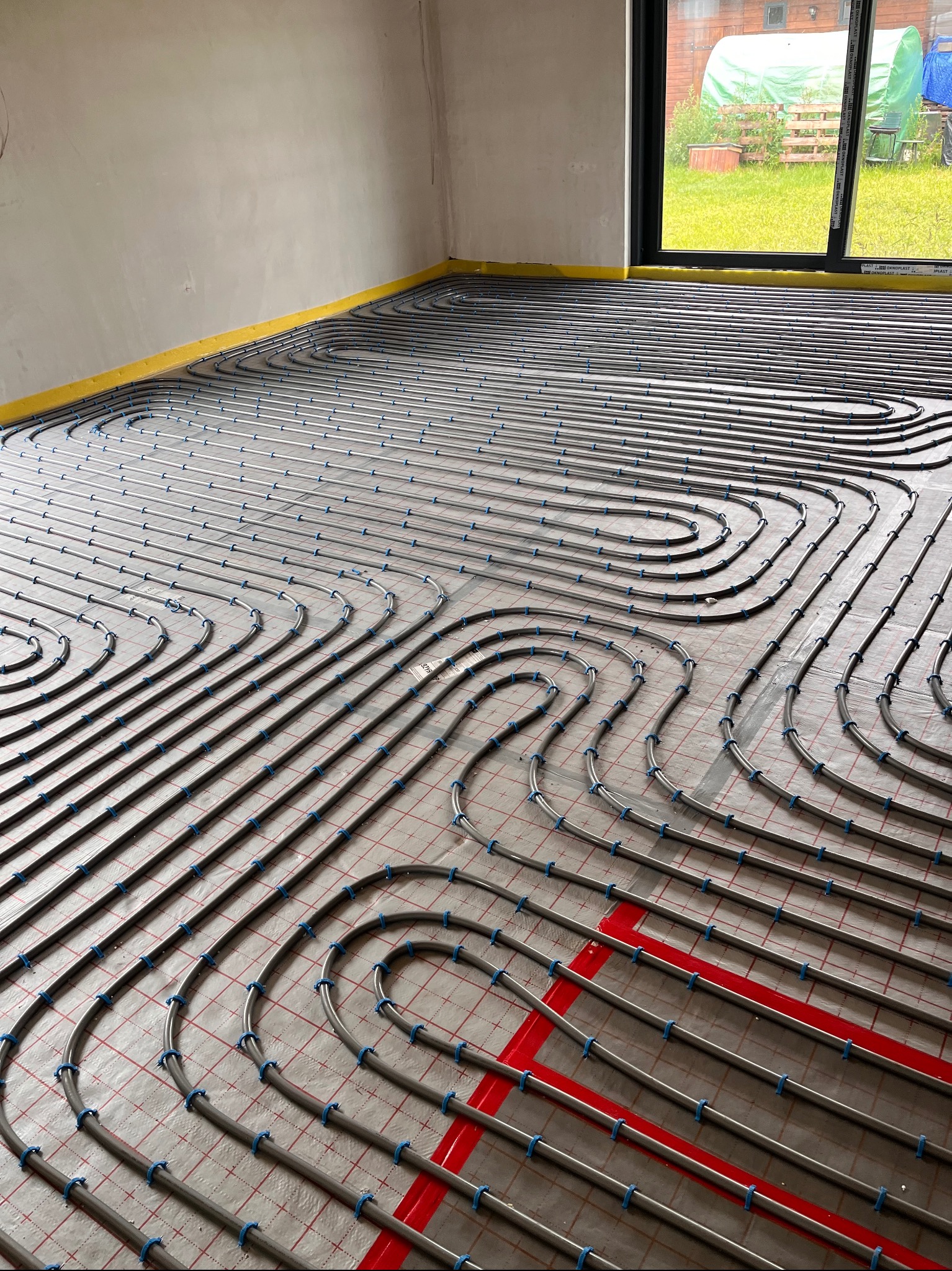 Ułożone rury ogrzewania podłogowego z niebieskimi uchwytami na folii z siatką, widok z góry, w pomieszczeniu w Śremie. Widoczne okno z zielonym ogrodem.