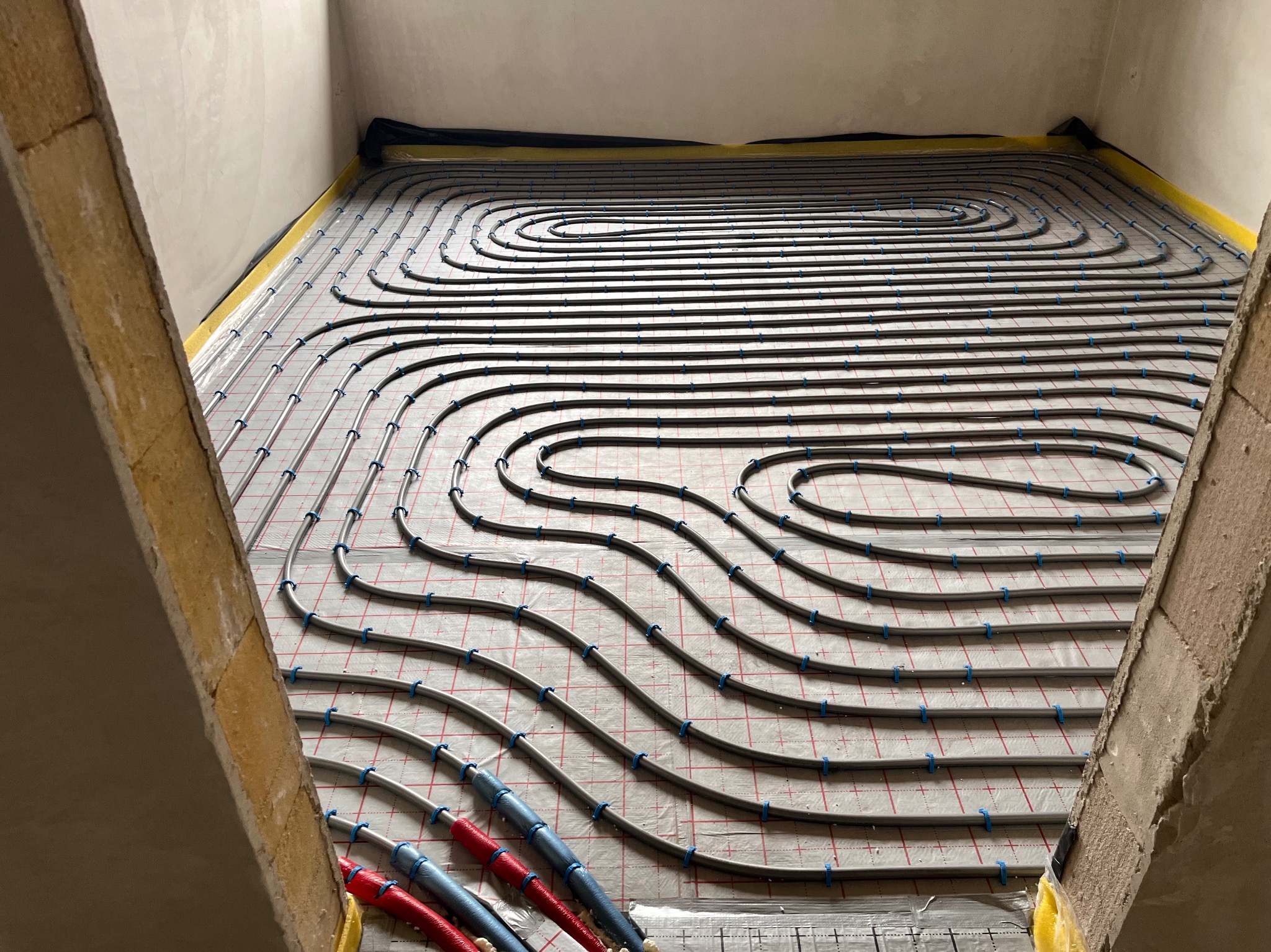 Ułożona instalacja ogrzewania podłogowego na folii z siatką, widoczne rury w równych odstępach, mocowane niebieskimi uchwytami. W tle ściany i izolacja.