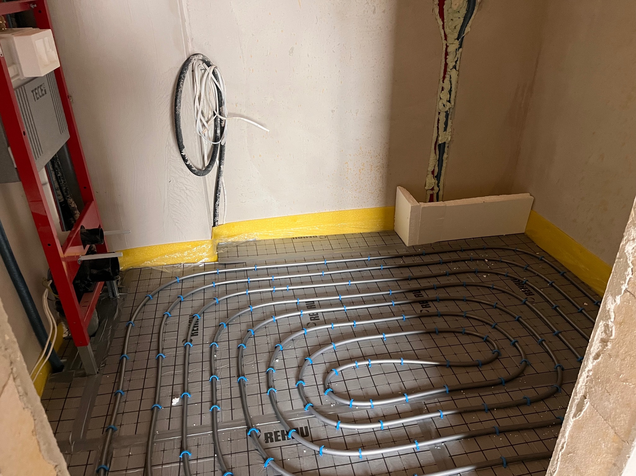 Szare rury ogrzewania podłogowego Rehau, zamocowane niebieskimi klipsami na czarnej macie, widoczne przyłącza i elementy instalacji. Żółta taśma izolacyjna wzdłuż ścian.