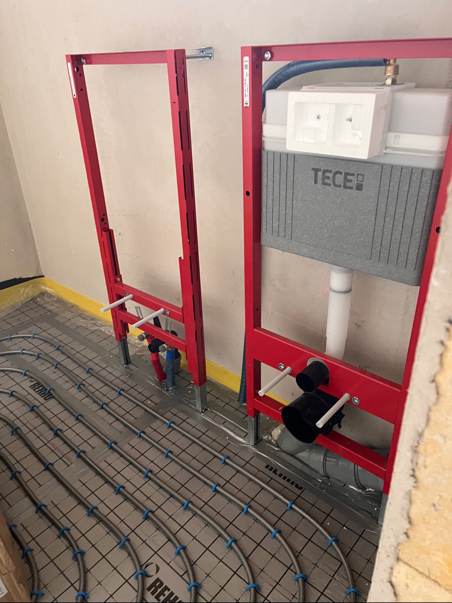 Czerwone stelaże podtynkowe TECE zamontowane na tle ułożonej instalacji ogrzewania podłogowego Rehau, widok z góry pod kątem. Etap prac instalacyjnych.