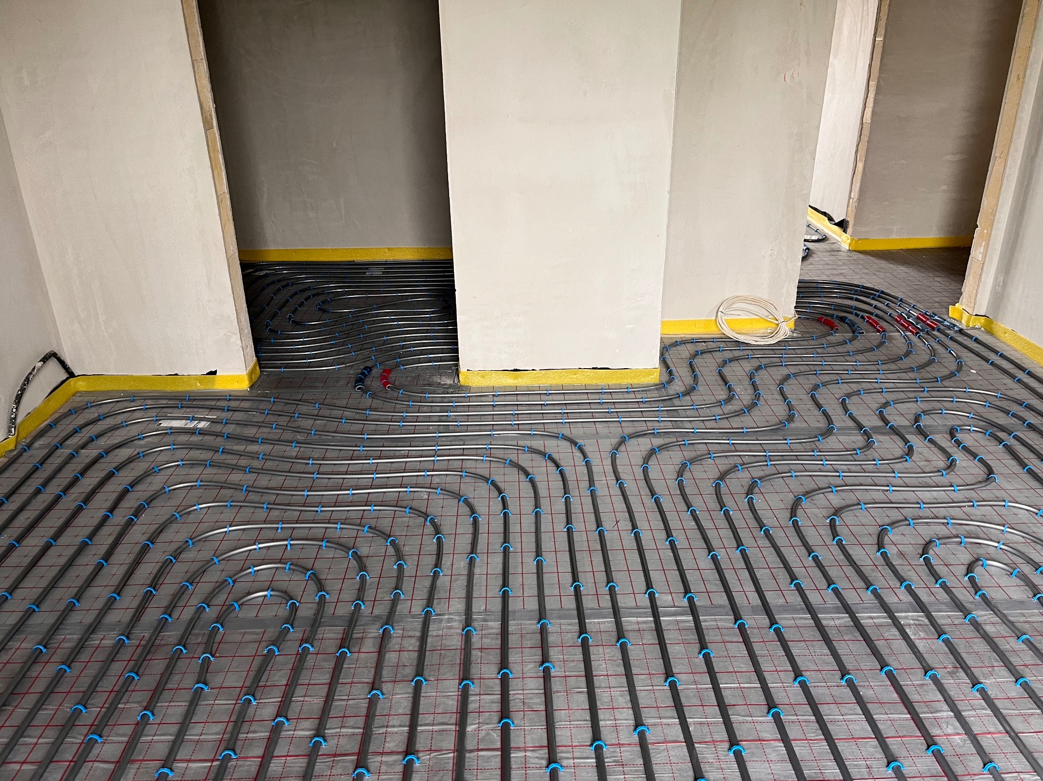 Szare rury ogrzewania podłogowego z niebieskimi mocowaniami ułożone na podkładzie z czerwoną siatką, zabezpieczone żółtą taśmą przy ścianach w surowym wnętrzu.