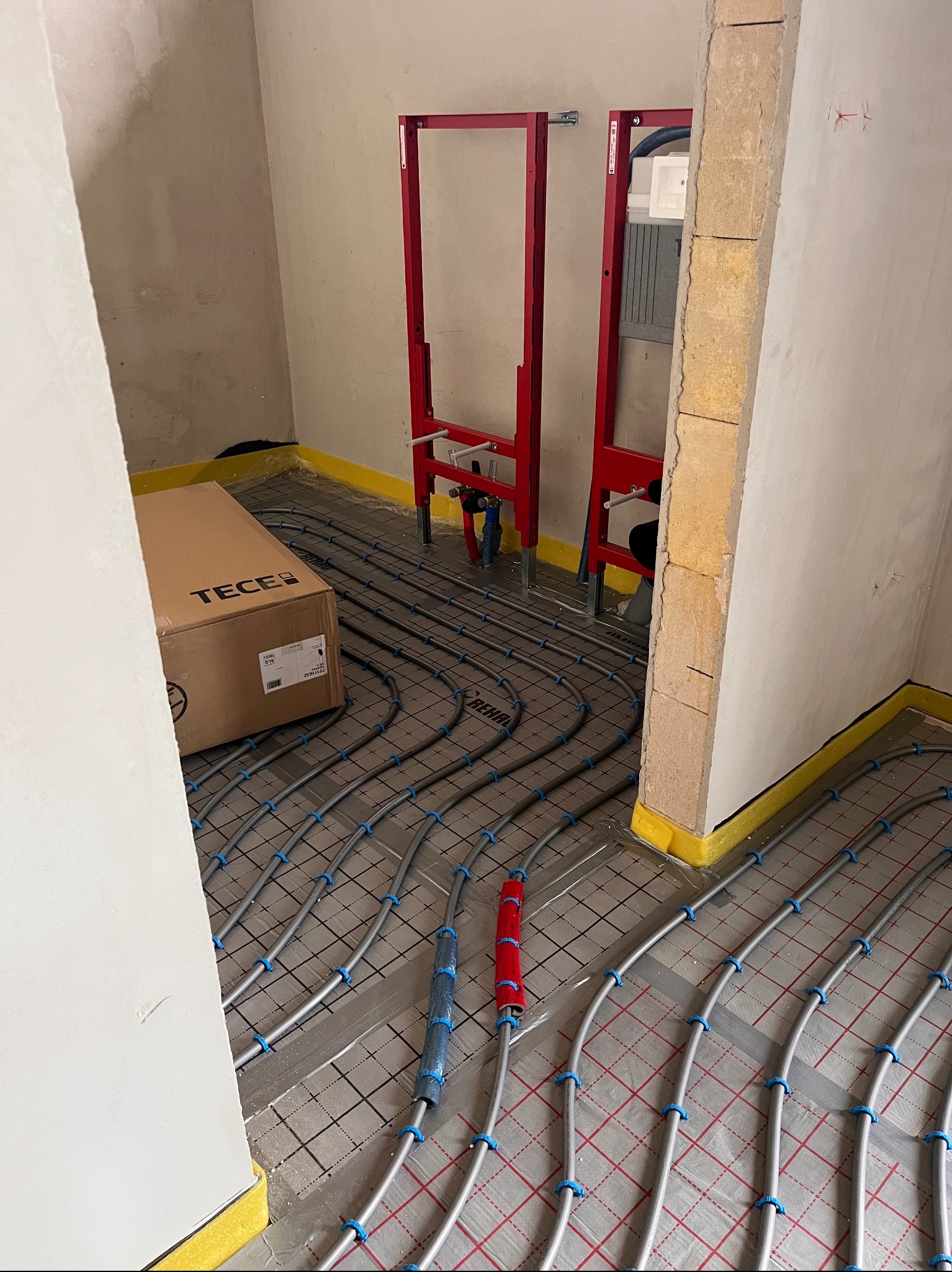 Szare rury ogrzewania podłogowego na macie z kratką, karton TECE i czerwone stelaże podtynkowe w surowym wnętrzu łazienki w trakcie instalacji.
