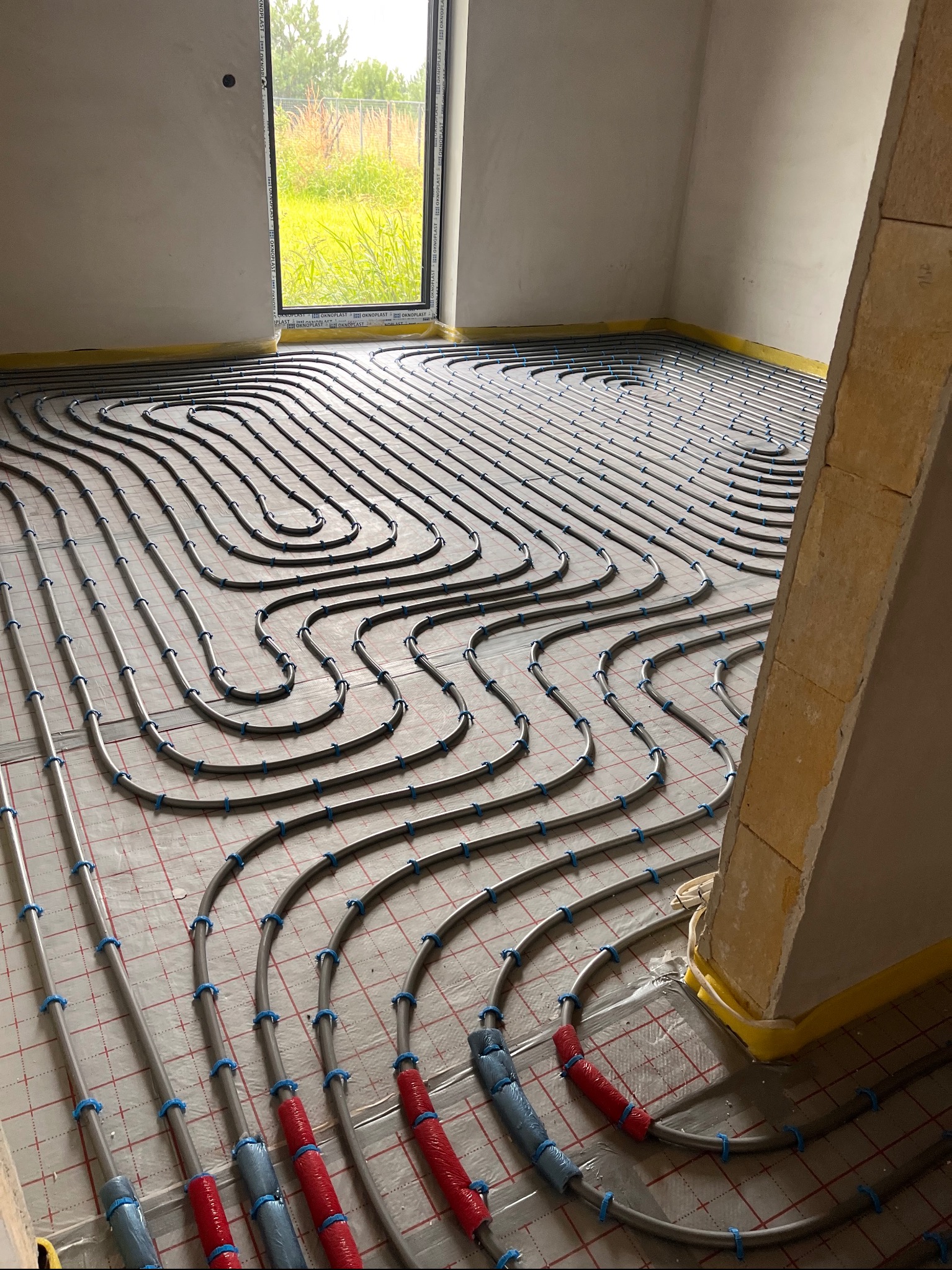 Szare rury ogrzewania podłogowego, przymocowane niebieskimi uchwytami do podłoża z siatką. Część rur zabezpieczona folią. W tle okno z widokiem na zieleń.