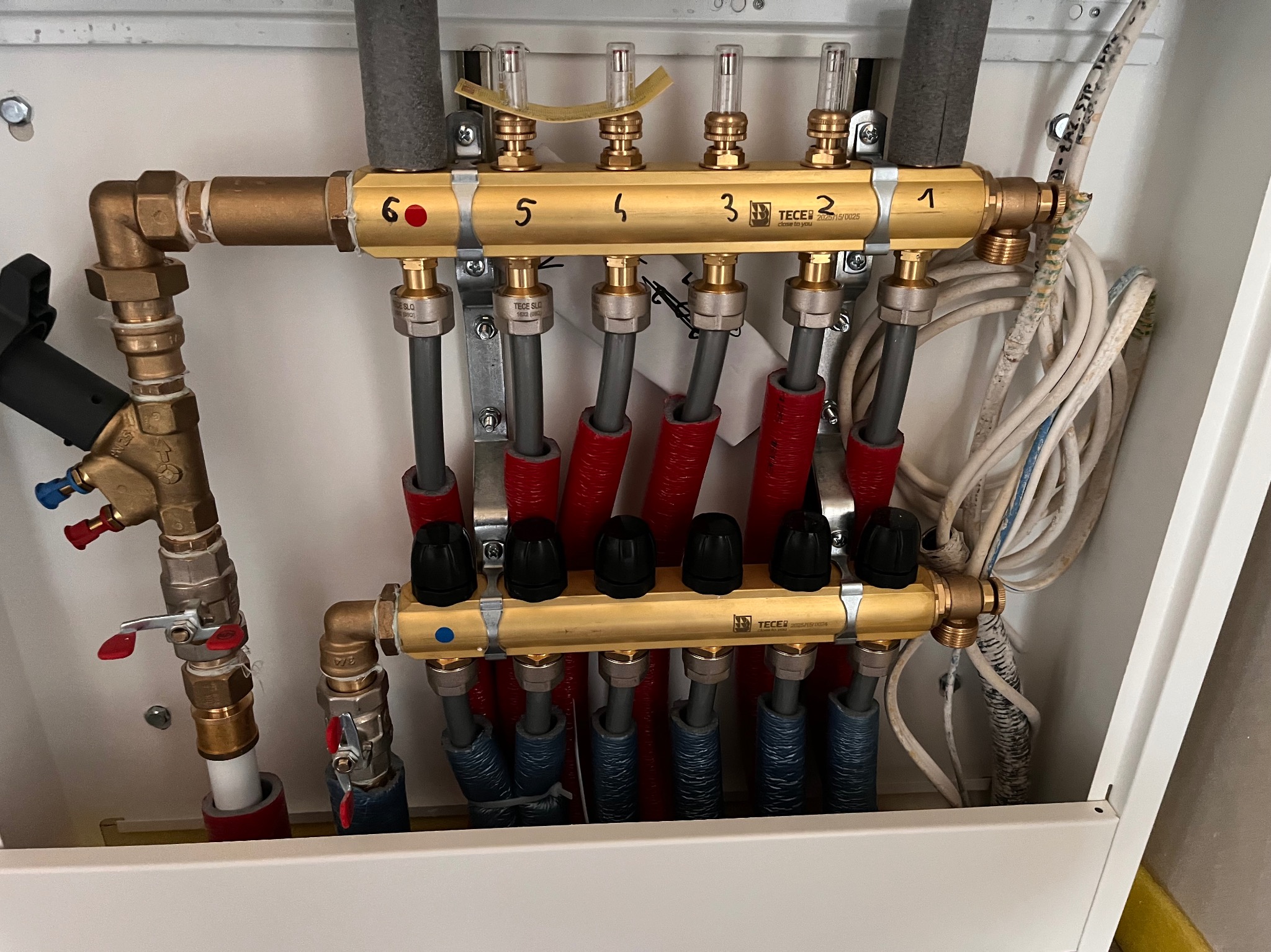 Rozdzielacz ogrzewania podłogowego TECE w szafce, z numerowanymi sekcjami (1-6) i rurami w izolacji czerwonej i niebieskiej. Widoczne zawory i przewody.
