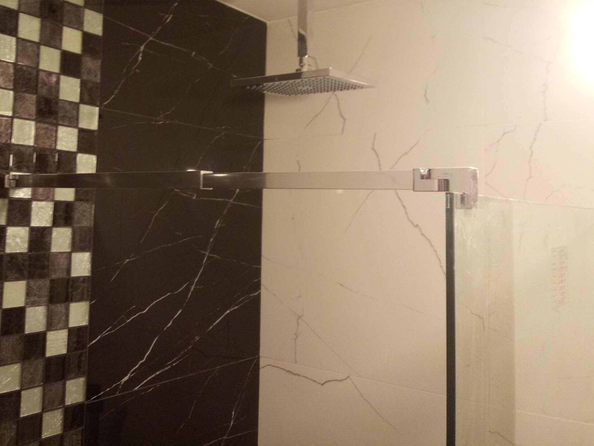 Kabina prysznicowa z deszczownicą i szklanymi drzwiami, wykończona mozaiką i płytkami imitującymi marmur w odcieniach bieli i czerni.