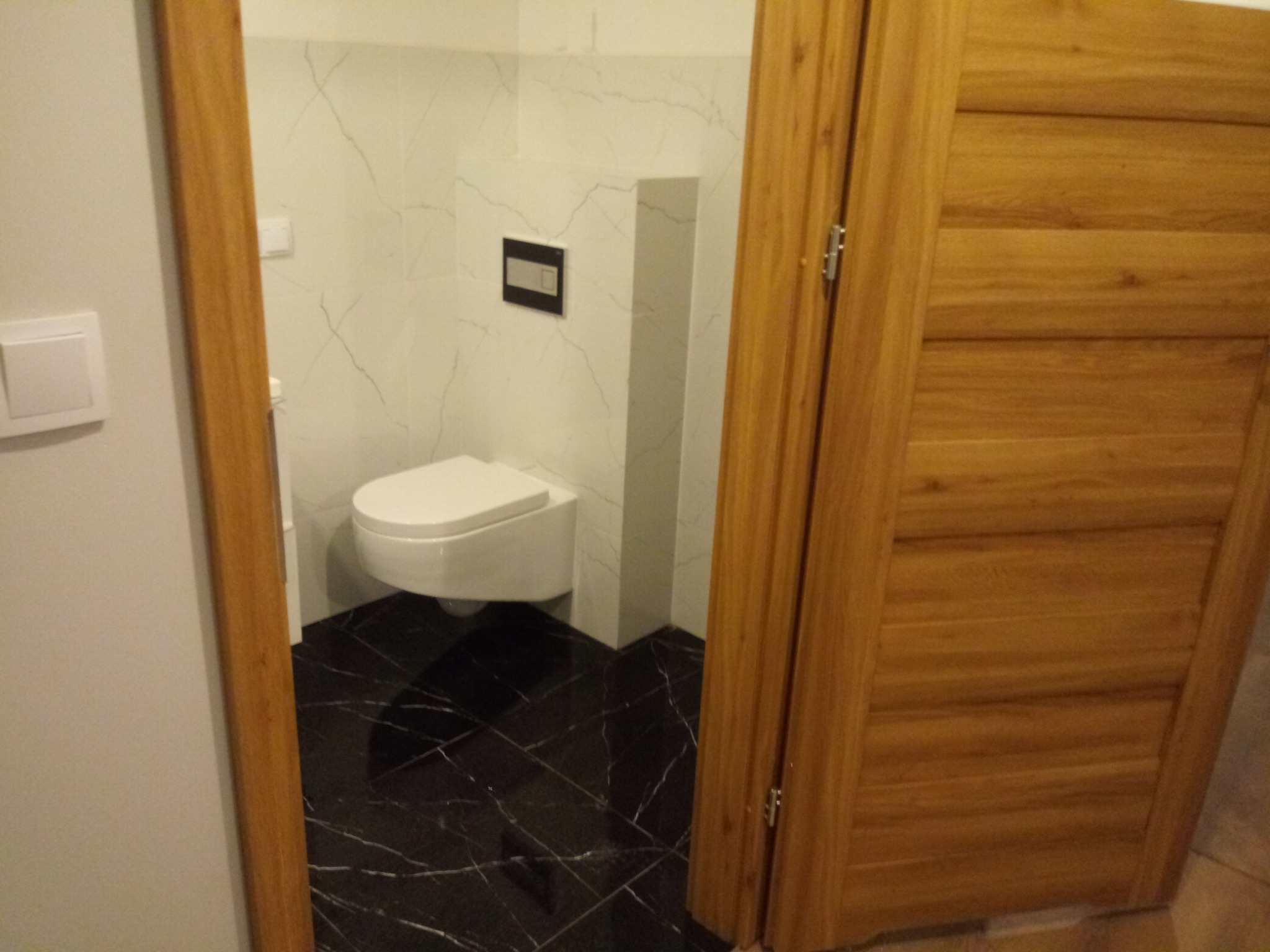 Nowoczesna toaleta podwieszana w łazience z czarną, marmurkową podłogą i białą okładziną ścienną imitującą marmur, widoczna przez uchylone drewniane drzwi.