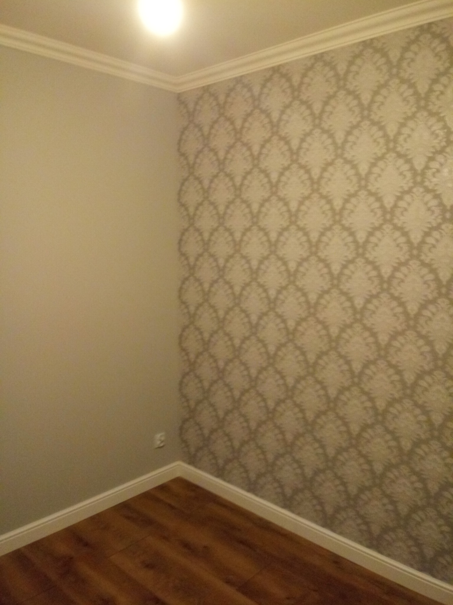 Fragment pokoju po remoncie: ściana z wzorzystą tapetą w odcieniach beżu i szarości, sąsiadująca z gładką, pomalowaną na szaro ścianą, widoczna listwa przypodłogowa i fragment podłogi z paneli...