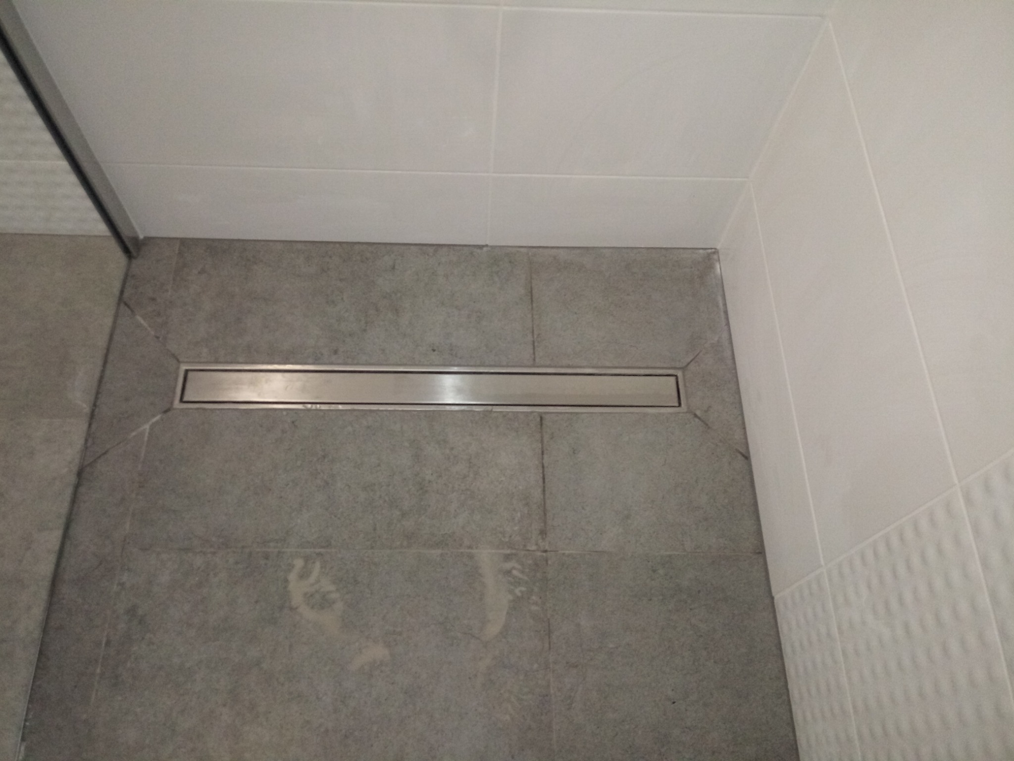 Szczegółowe ujęcie odpływu liniowego w kabinie prysznicowej z szarymi płytkami podłogowymi i białymi płytkami ściennymi z delikatną strukturą. Widoczne precyzyjne wykończenie i kąt nachylenia...