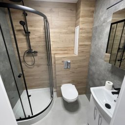 M-Bud Maciej Lorenc - Nowoczesna łazienka po remoncie: kabina prysznicowa z czarnymi ramami, toaleta, umywalka z szafką. Ściany wyłożone płytkami imitującymi drewno i szarym betonem.