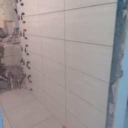 USŁUGI BUDOWLANE MATEUSZ PIĘTKA - Świeżo ułożone, jasne płytki ceramiczne na ścianie i podłodze w trakcie remontu łazienki, z widocznymi klipsami poziomującymi i fragmentami surowego muru.