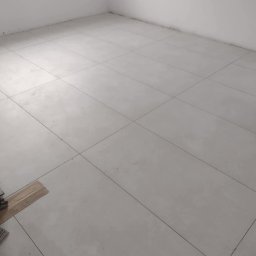 USŁUGI BUDOWLANE MATEUSZ PIĘTKA - Jasne, kwadratowe płytki podłogowe ułożone w pustym, białym pokoju. Widoczne deski na podłodze w lewym dolnym rogu.