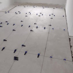 USŁUGI BUDOWLANE MATEUSZ PIĘTKA - Układanie jasnych płytek podłogowych z użyciem niebieskich klipsów poziomujących, widoczne fugi i niezagruntowane ściany w tle.