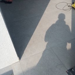 USŁUGI BUDOWLANE MATEUSZ PIĘTKA - Ułożona szara, matowa podłoga z płytek gresowych na zewnątrz, cień sylwetki osoby i kabel od urządzenia, w tle widoczne klapki.