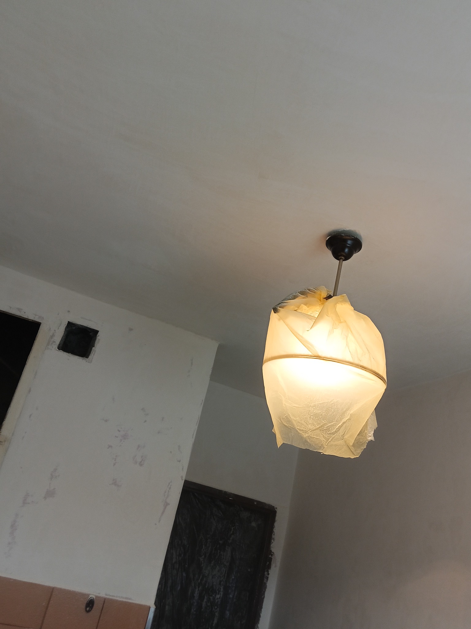 Lampa sufitowa zabezpieczona folią malarską podczas prac remontowych, widoczne ślady po malowaniu na ścianach i suficie.