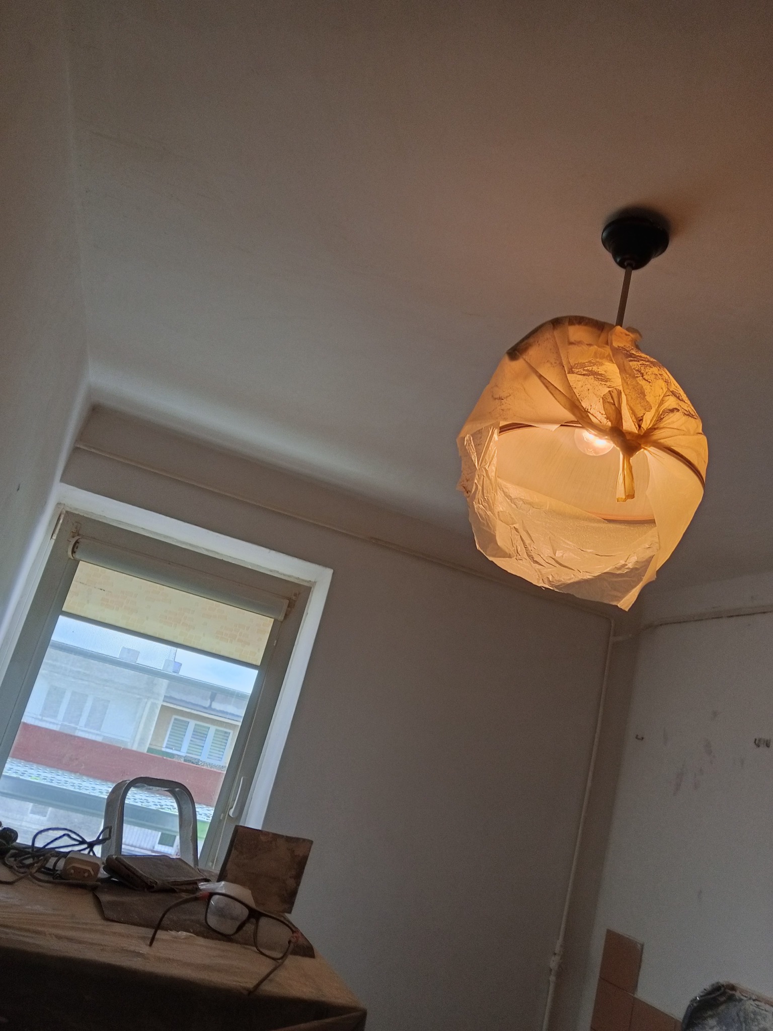Lampa zabezpieczona folią malarską podczas prac remontowych, widoczne narzędzia na stole i niedokończone ściany.