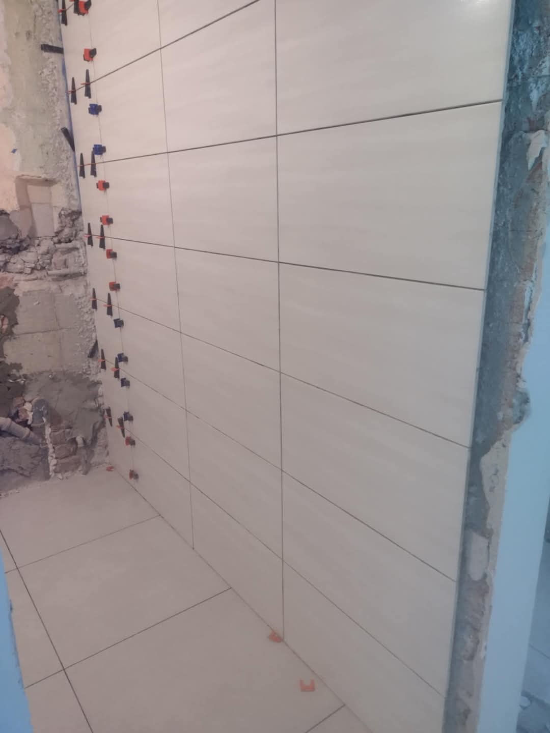 Świeżo ułożone, jasne płytki ceramiczne na ścianie i podłodze w trakcie remontu łazienki, z widocznymi klipsami poziomującymi i fragmentami surowego muru.