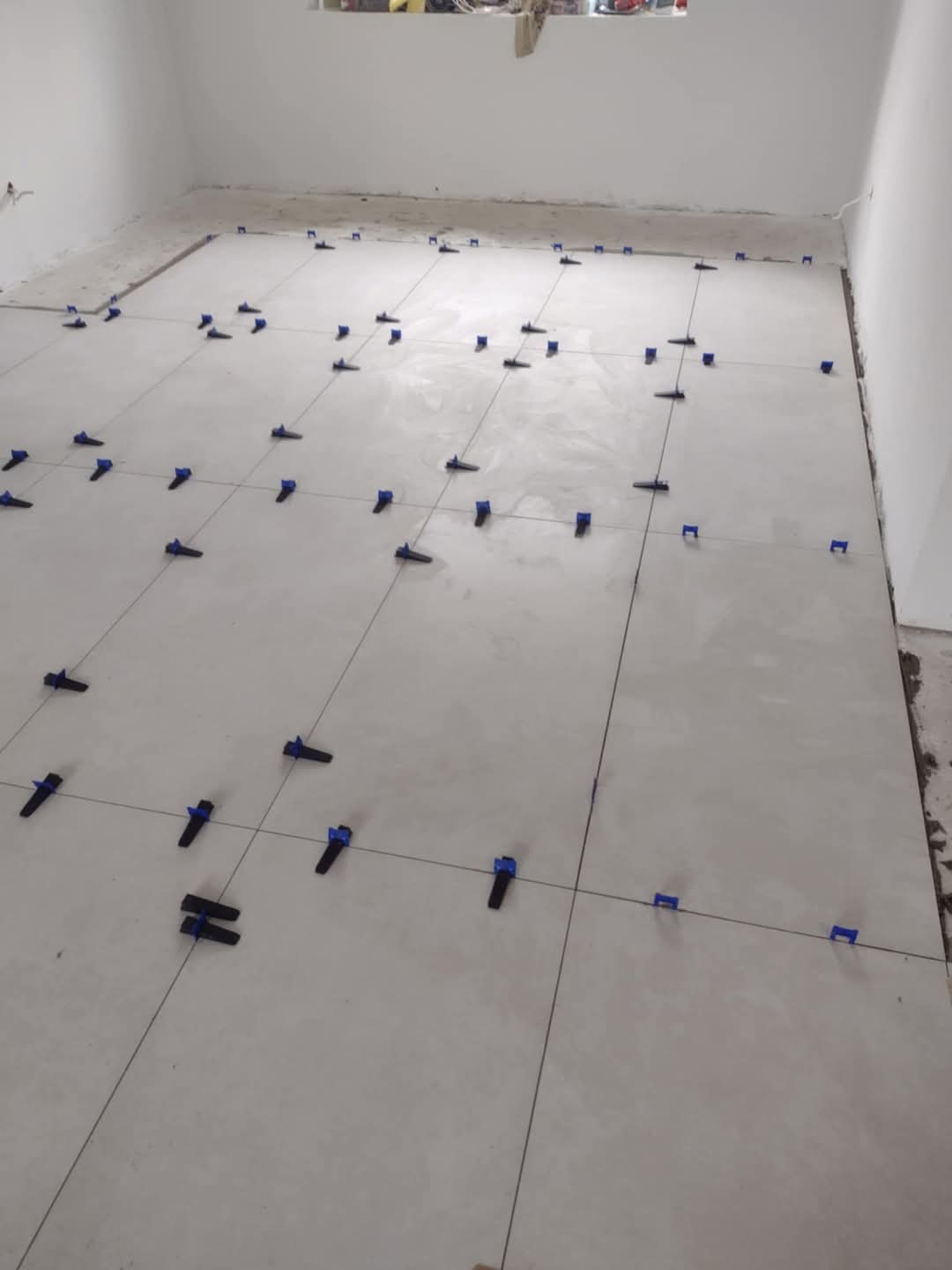 Układanie jasnych płytek podłogowych z użyciem niebieskich klipsów poziomujących, widoczne fugi i niezagruntowane ściany w tle.