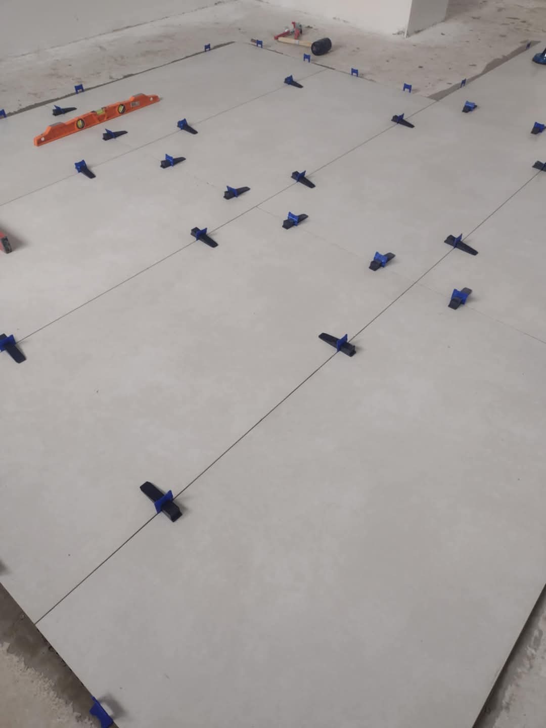 Układanie jasnych, matowych płytek podłogowych z użyciem systemu poziomowania, widoczna poziomica i wyznaczone linie.