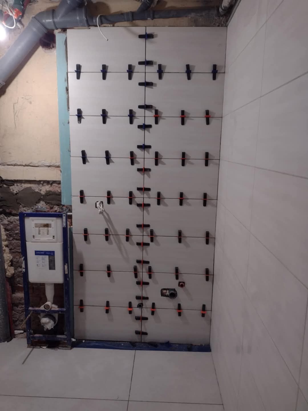 Montaż białych płytek ceramicznych na ścianie w łazience, z użyciem krzyżyków dystansowych, widoczny stelaż podtynkowy WC.