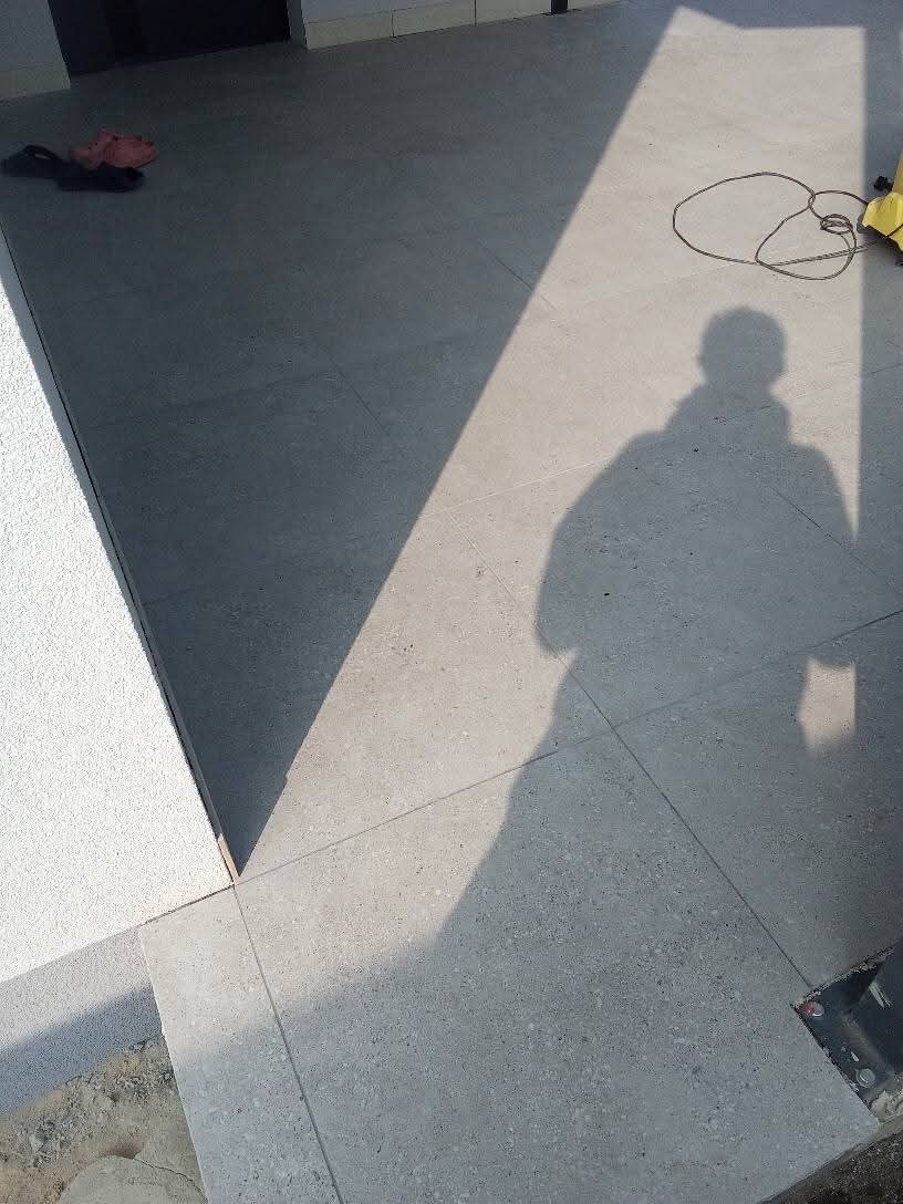 Ułożona szara, matowa podłoga z płytek gresowych na zewnątrz, cień sylwetki osoby i kabel od urządzenia, w tle widoczne klapki.
