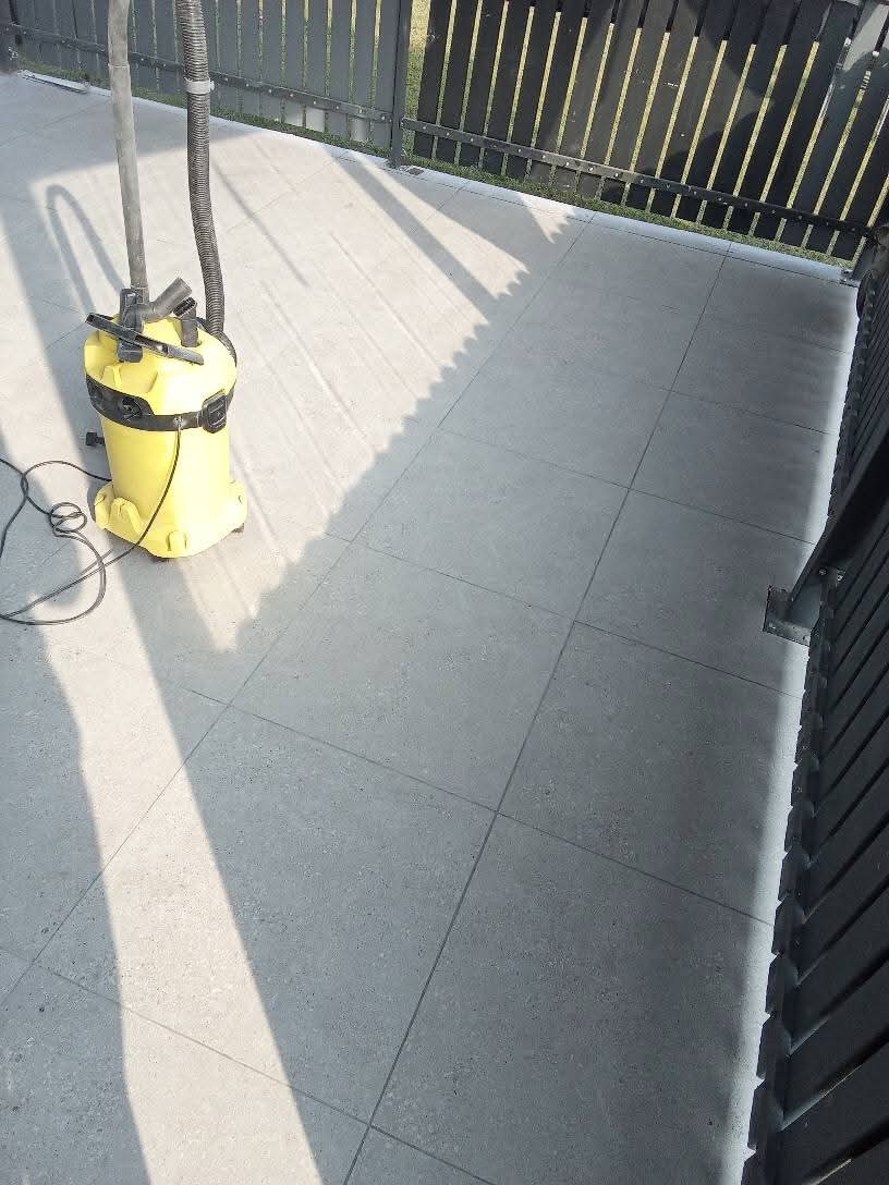 Szare płytki podłogowe na balkonie po czyszczeniu, z widocznym żółtym odkurzaczem przemysłowym i fragmentem czarnego ogrodzenia w tle.