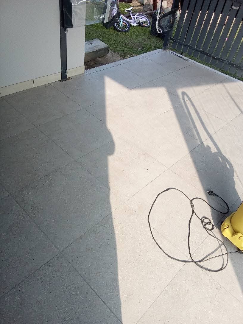 Szare płytki podłogowe na zewnątrz, częściowo zacienione, z widocznym kablem i fragmentem żółtej myjki ciśnieniowej; w tle fragment trawnika z rowerem.