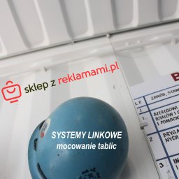 sklepzreklamami - Niebieski hełm z widocznymi systemami linkowymi mocowania tablic, umieszczony obok tablicy z listą zawodów i stanowisk wewnątrz białego kontenera.