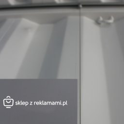 sklepzreklamami - Szary panel z białym napisem 'sklep z reklamami.pl' oraz 'Mocowanie tablic Systemy Linkowe', zamocowany na białej ścianie za pomocą metalowych linek i uchwytów.