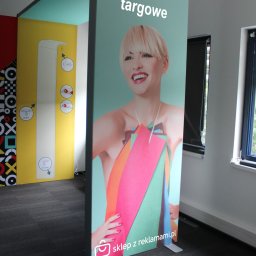 sklepzreklamami - Stoisko targowe z uśmiechniętą blondynką i kolorowymi elementami graficznymi, widoczne logo 'sklep z reklamami.pl' w dolnej części, w tle fragmenty innych stoisk i okno.