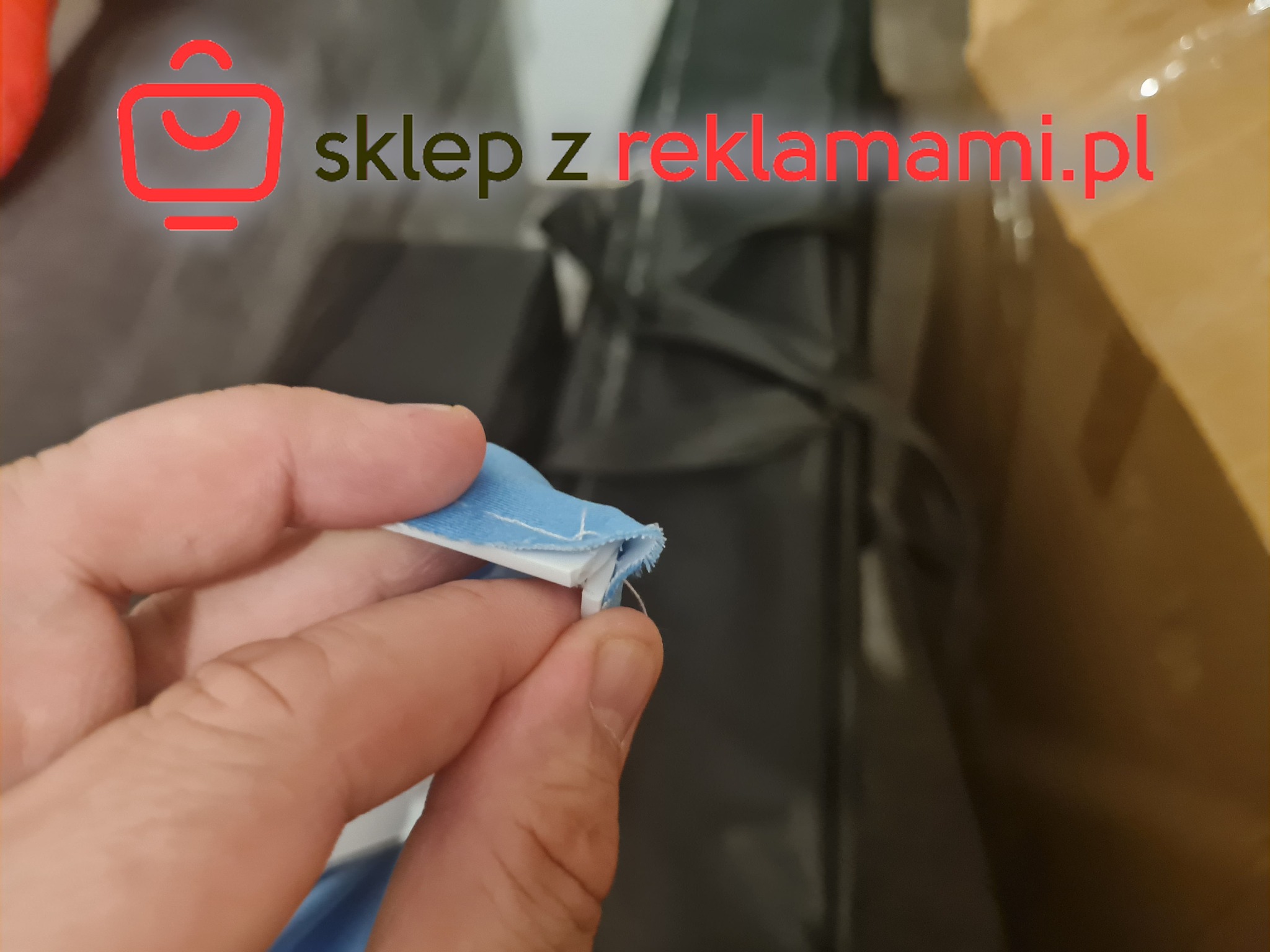 Detal dłoni trzymającej fragment niebieskiej tkaniny z białym brzegiem na tle pudeł i logo sklepu z reklamami