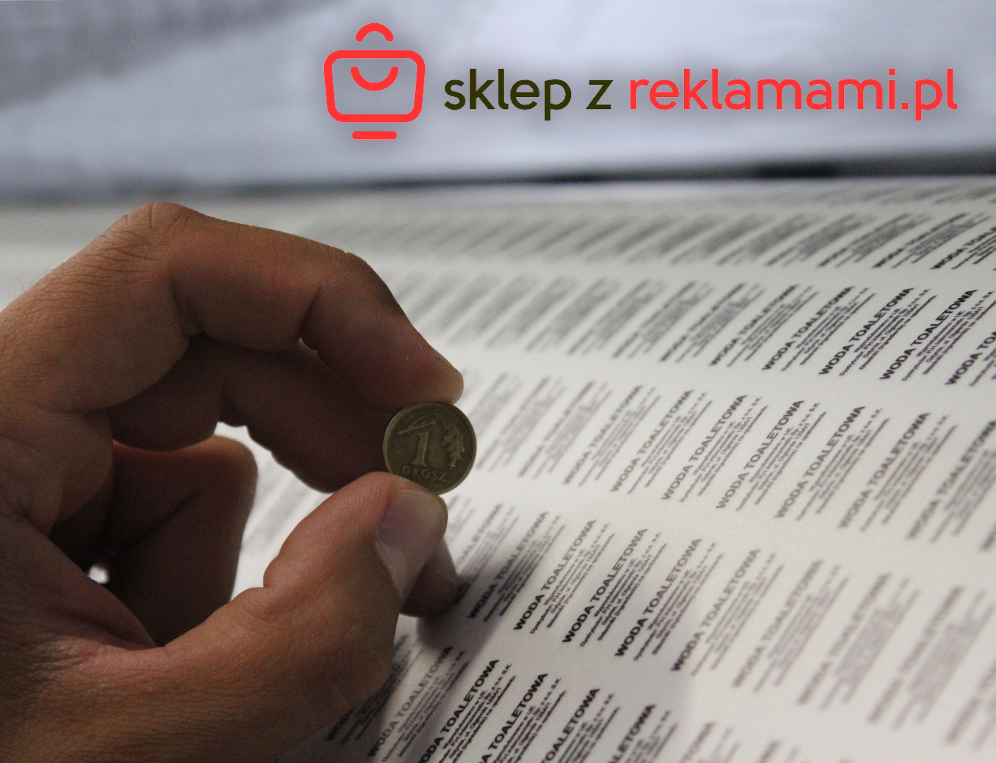 Dłoń trzymająca jednogroszówkę na tle arkusza z powtarzalnymi, czarnymi napisami 'WODA TOALETOWA', przykładowa realizacja druku etykiet.