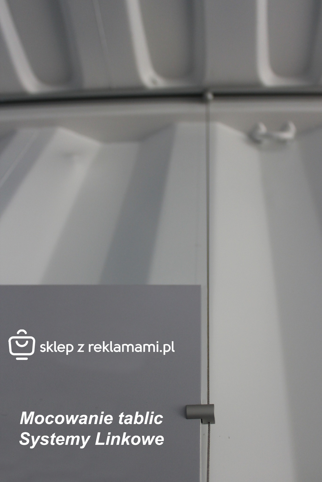 Szary panel z białym napisem 'sklep z reklamami.pl' oraz 'Mocowanie tablic Systemy Linkowe', zamocowany na białej ścianie za pomocą metalowych linek i uchwytów.