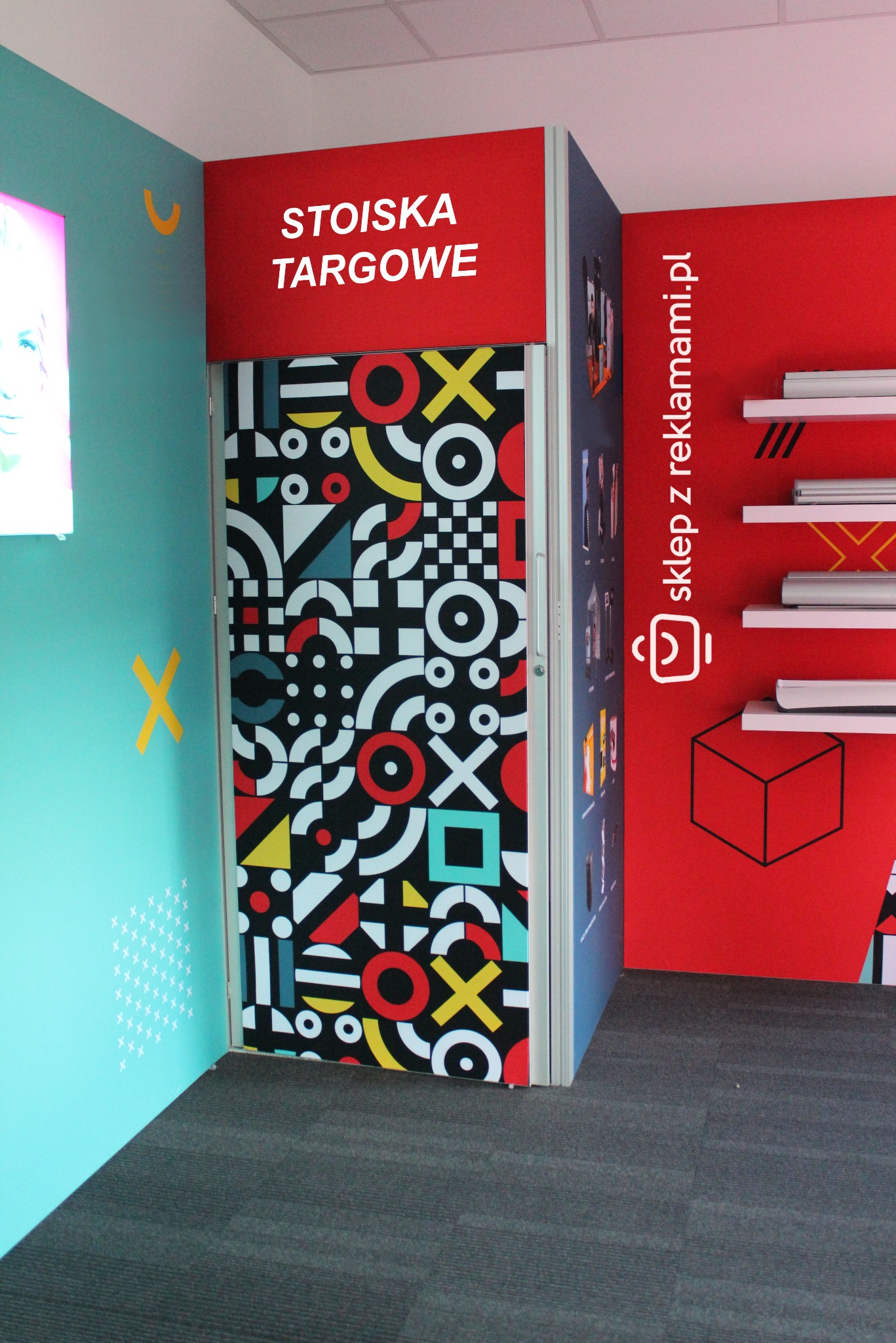 Stoisko targowe z geometrycznym wzorem na drzwiach, czerwonym panelem z napisem 'STOISKA TARGOWE' i elementami graficznymi sklepu z reklamami na czerwonej ścianie.