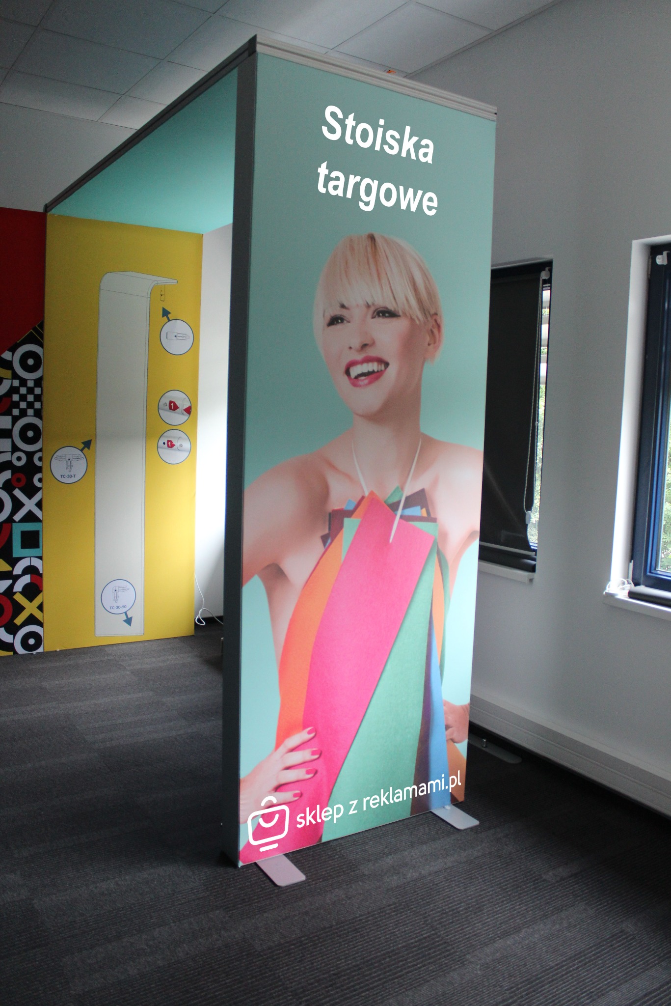 Stoisko targowe z uśmiechniętą blondynką i kolorowymi elementami graficznymi, widoczne logo 'sklep z reklamami.pl' w dolnej części, w tle fragmenty innych stoisk i okno.