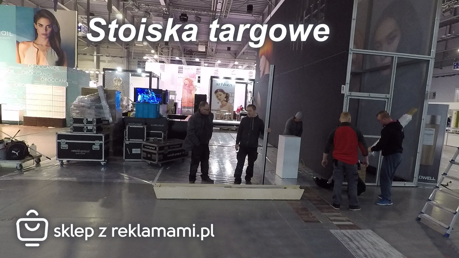 Montaż stoiska targowego: ekipa budowlana w trakcie pracy, widoczne elementy konstrukcyjne, plakaty reklamowe i skrzynie transportowe w hali wystawienniczej.
