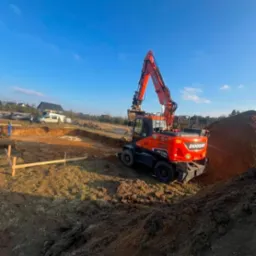 Pomarańczowa koparko-ładowarka Doosan DX170W-5 na placu budowy, widok z tyłu, świeżo wykopany rów, drewniane szalunki w tle, błękitne niebo.