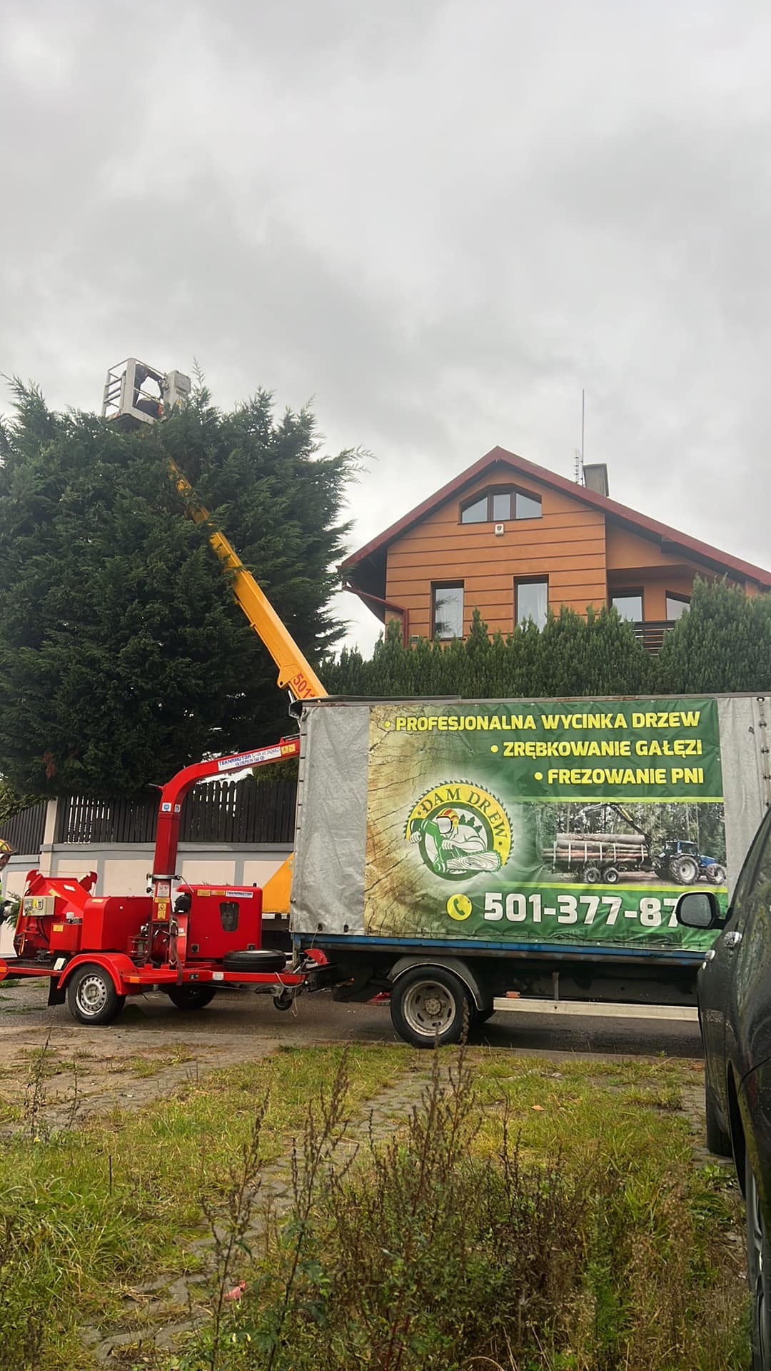 Wycinka drzew z podnośnika koszowego przy domu. Na aucie reklama firmy z numerem telefonu. Czerwony rębak do gałęzi. Szare niebo.