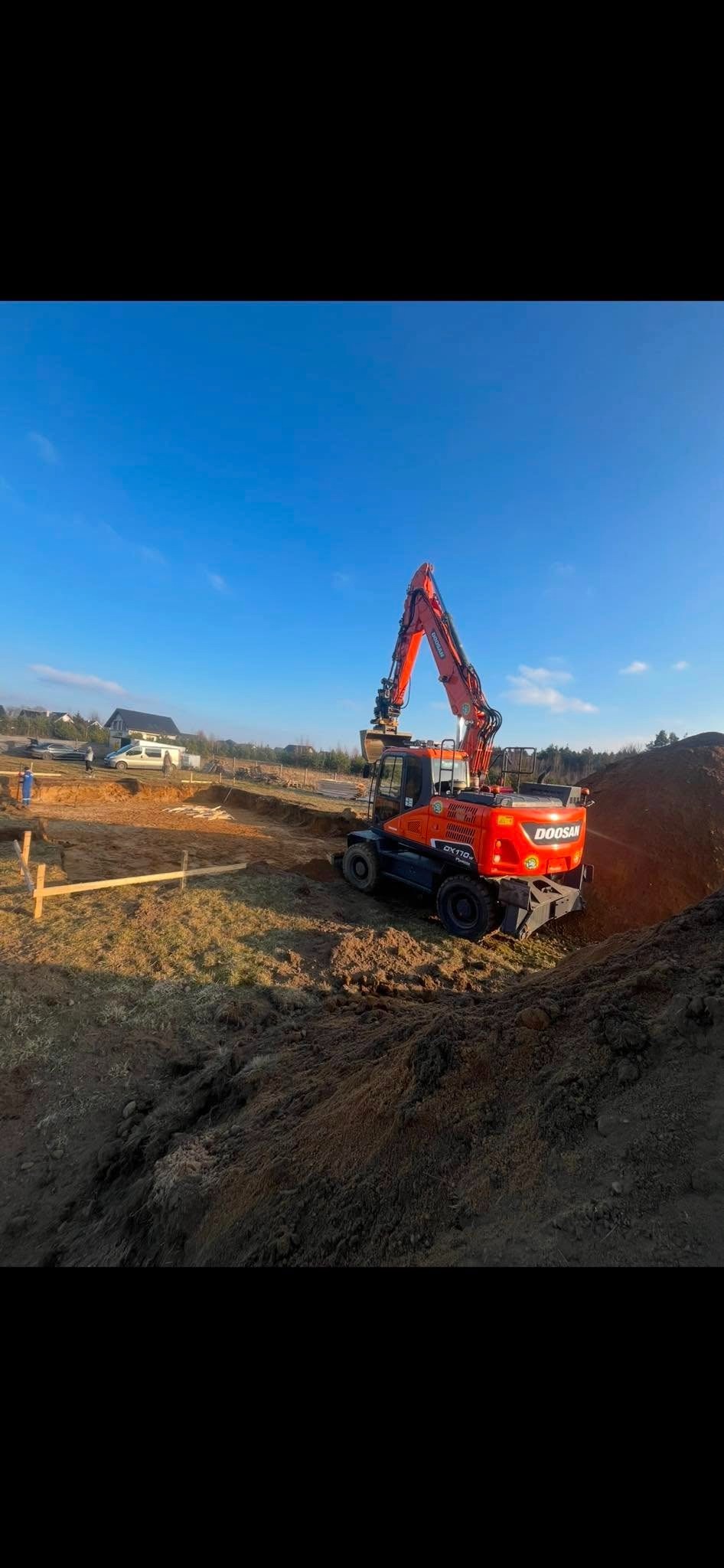 Pomarańczowa koparko-ładowarka Doosan DX170W-5 na placu budowy, widok z tyłu, świeżo wykopany rów, drewniane szalunki w tle, błękitne niebo.