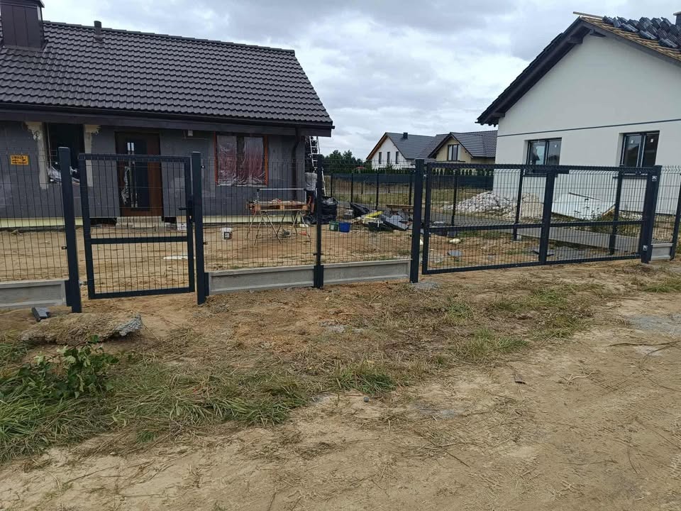 Nowoczesne ogrodzenie panelowe w kolorze grafitowym, z betonową podmurówką, otaczające posesję z dwoma domami w tle. Widoczny otwarty wjazd.
