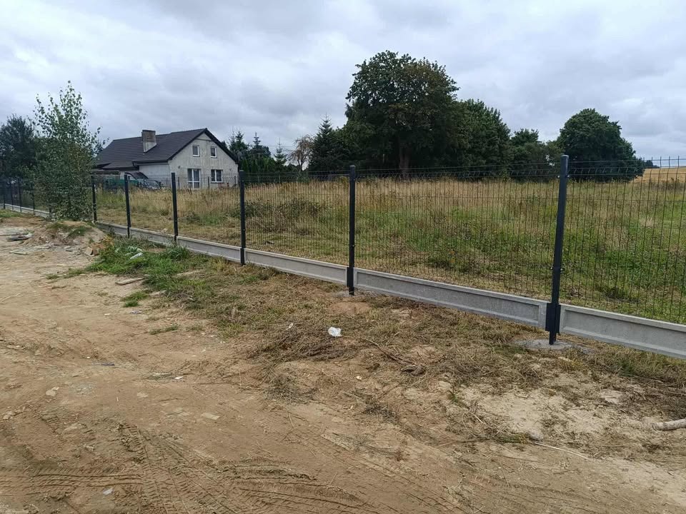 Nowoczesne ogrodzenie panelowe z podmurówką betonową na nierównym terenie, dom w tle. Widoczne ślady opon na ziemi. Pochmurne niebo.