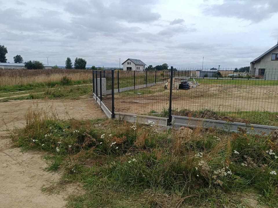 Częściowo ukończone ogrodzenie panelowe z podmurówką betonową na nierównym terenie z widokiem na domy w tle, pochmurne niebo.