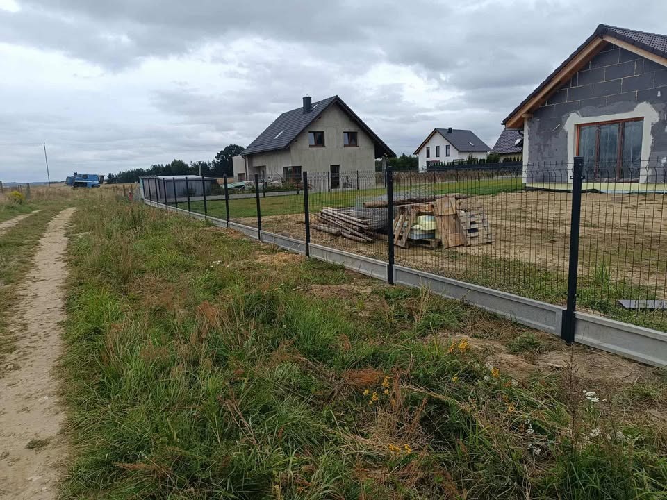 Nowe ogrodzenie panelowe z podmurówką, widok z zewnątrz posesji. Na drugim planie domy w różnym stanie wykończenia i pochmurne niebo.