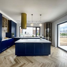 Marta Kieleczawa Project Solutions - Nowoczesna kuchnia w odcieniach granatu z wyspą i złotymi akcentami. Parkiet ułożony w jodełkę, duże okno z widokiem na zewnątrz. Minimalistyczny design.