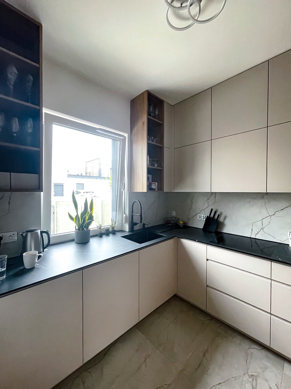 Jasna kuchnia z czarnym blatem i marmurowym backsplash. Nowoczesne szafki bez uchwytów, czarny zlew pod oknem z roślinami. Minimalistyczny design z elementami drewna.