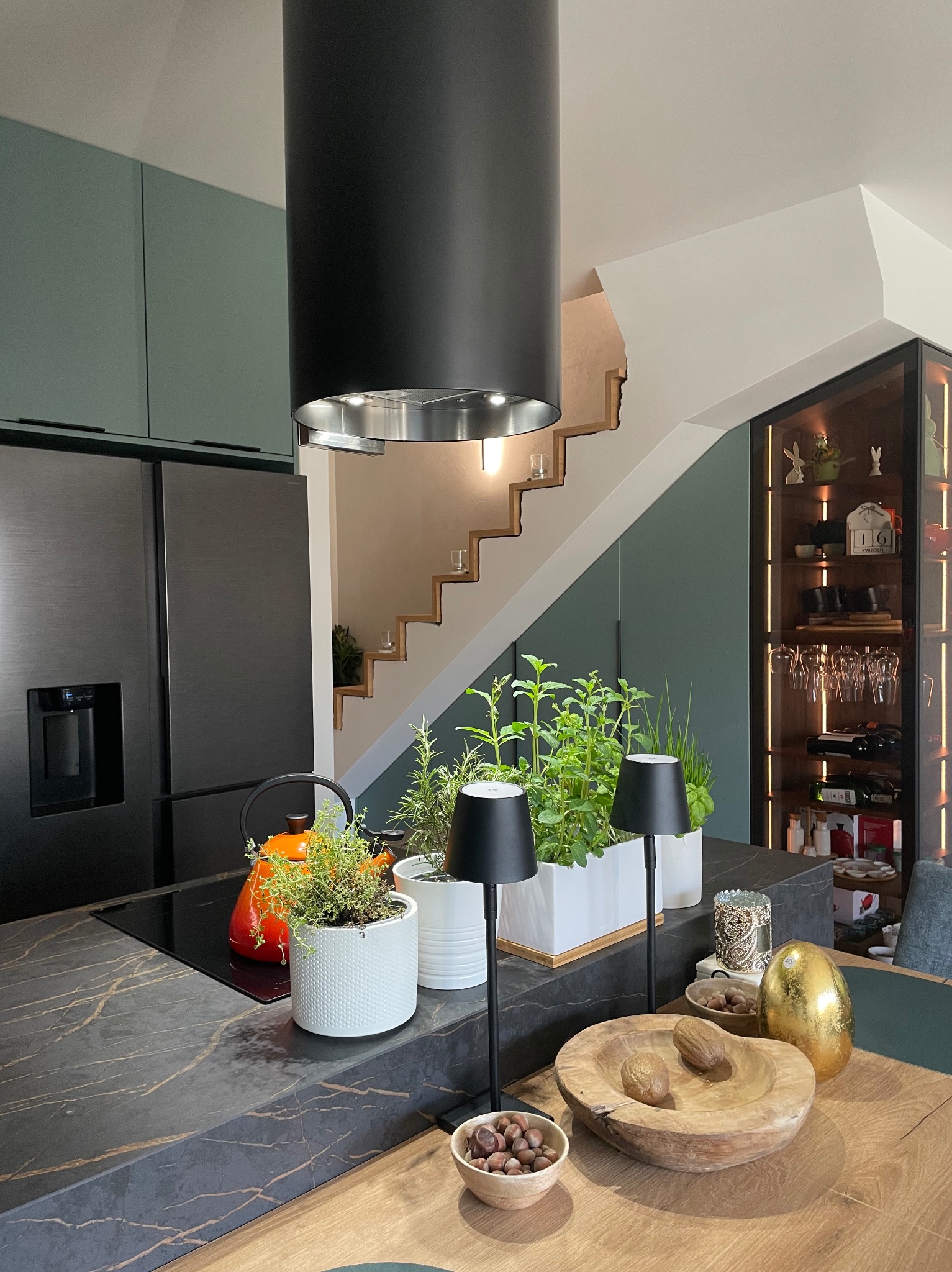 Nowoczesna kuchnia z czarnym okapem cylindrycznym, ziołami w doniczkach i drewnianą misą na blacie. Widoczne schody i przeszklona witryna z oświetleniem.