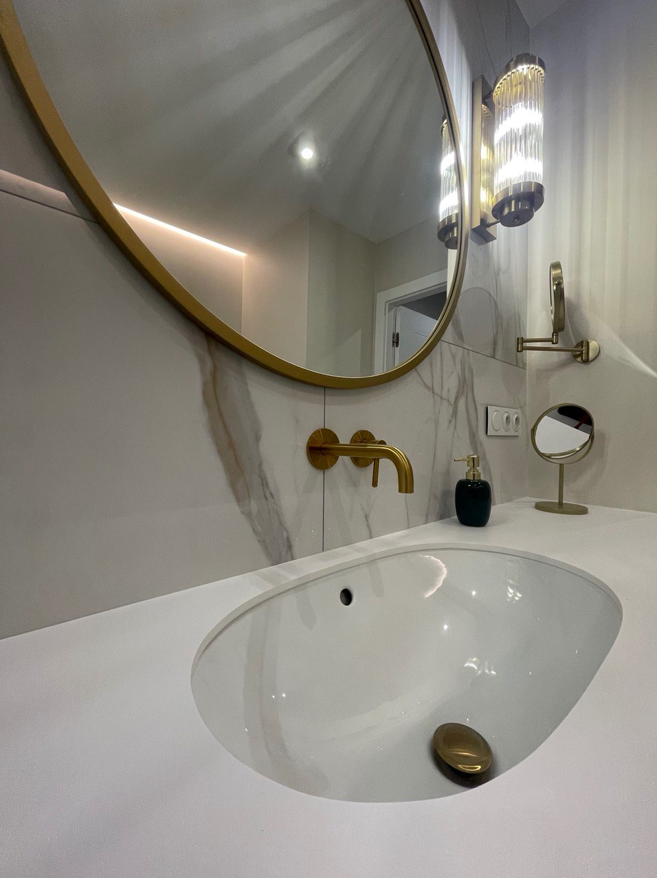 Biała umywalka z baterią w kolorze złota, okrągłe lustro z podświetleniem, marmurowe płytki i designerskie lampy w łazience.