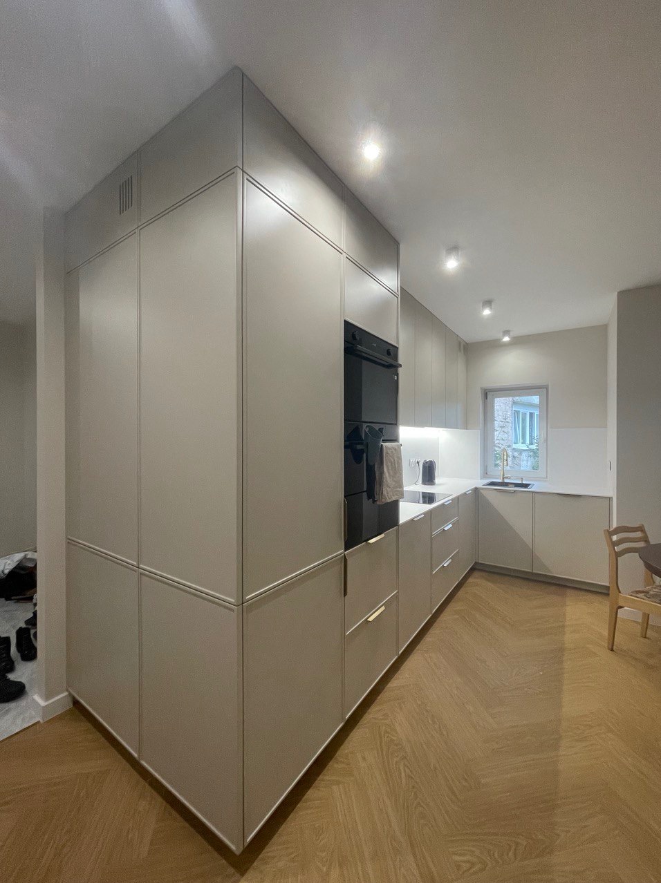 Nowoczesna kuchnia w odcieniach szarości z drewnianą podłogą w jodełkę. Zabudowa kuchenna z piekarnikiem i oknem w tle. Minimalistyczny design.