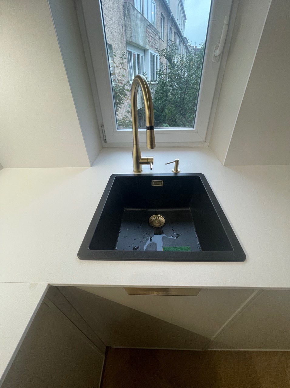 Czarny zlew granitowy z złotą baterią i dozownikiem mydła przy oknie dachowym z widokiem na budynek i drzewa, biały blat, nowoczesny design.