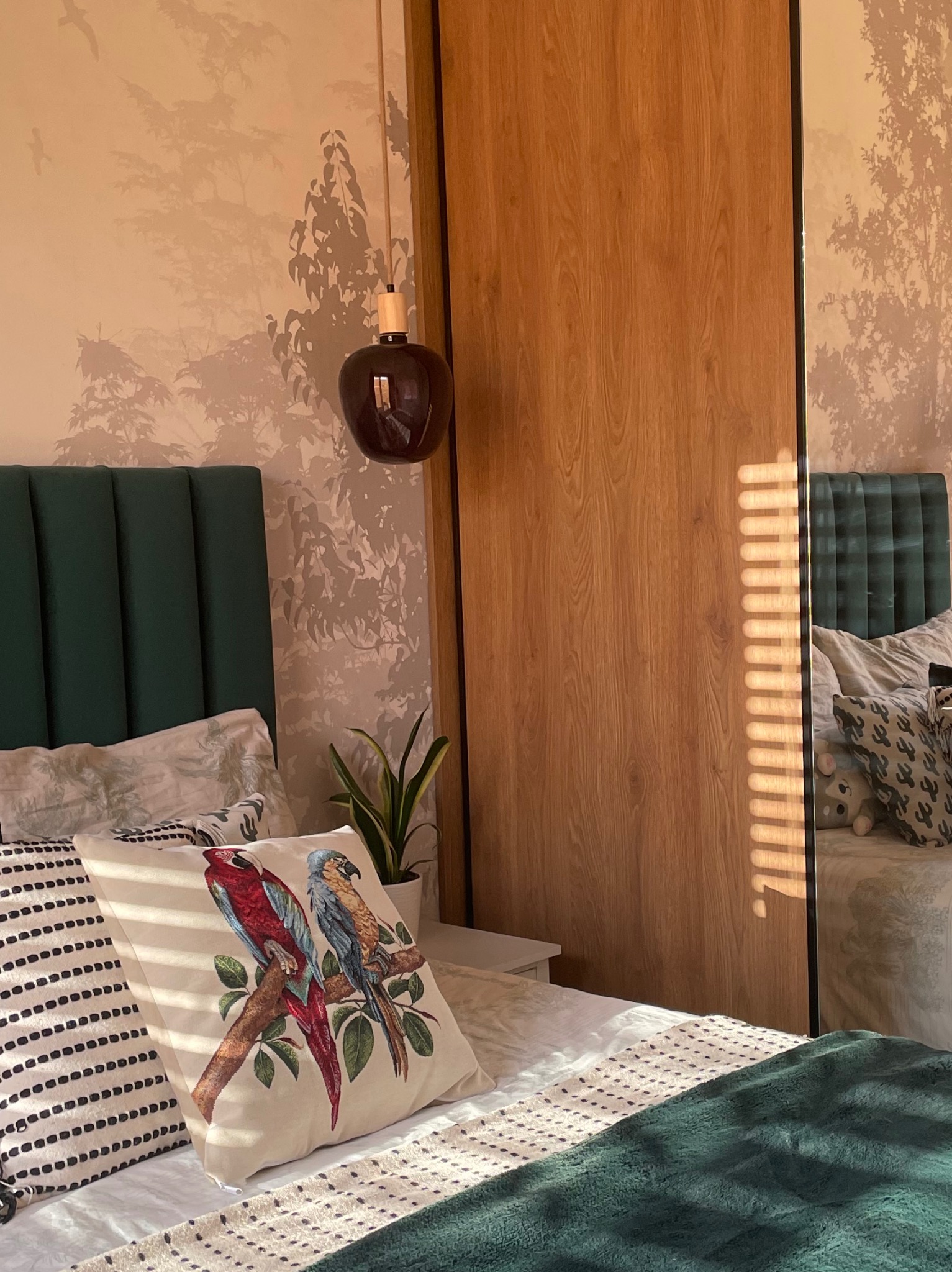 Sypialnia z zielonym zagłówkiem, poduszką z papugami i lampą wiszącą. Na ścianie tapeta z motywem drzew. W tle szafa z lustrem odbijającym fragment pokoju.